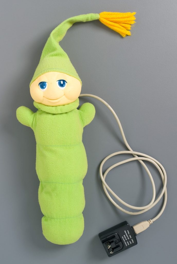 The Glo Worm is a soft plush toy in a bright green colour. The face is plastic and the body, arms and nightcap are green fabric. The nightcap has an orange tassel. Inside the body is a child-proof battery box and wire which activates the light which makes the face glow. When the body is pressed the light is turned on.